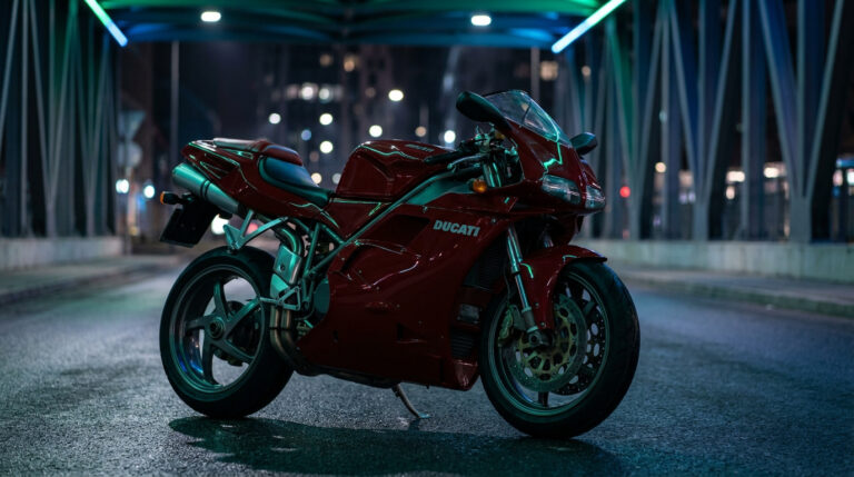 Moto Ducati Matrix rouge rutilante, stationnée de nuit sur une rue mouillée. Des néons verts et bleus illuminent l'arrière-plan urbain et un pont.