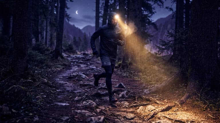 Homme en tenue de trail courant dans une forêt sombre la nuit, éclairé par sa lampe frontale. Une lune est visible.